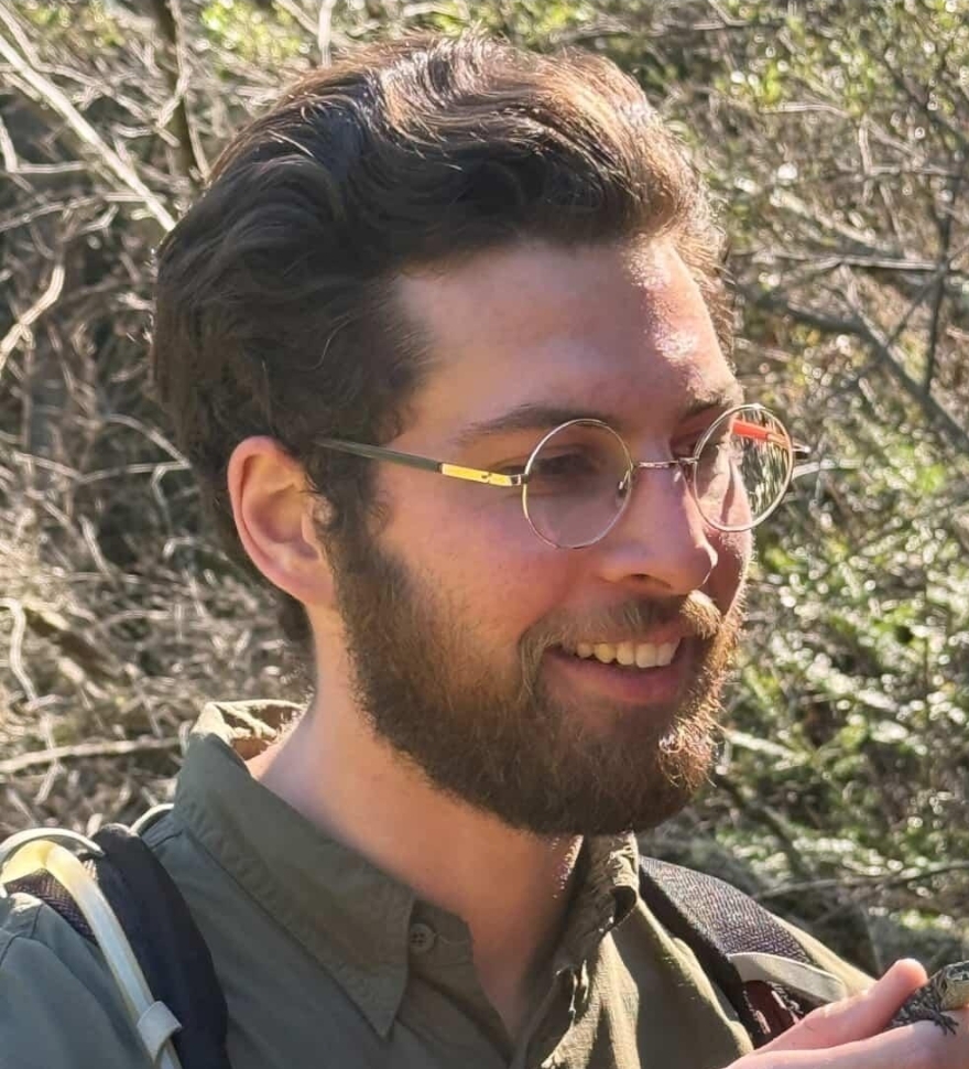 Nick smiling outdoors in dappled sunlight, backpack on, in the field